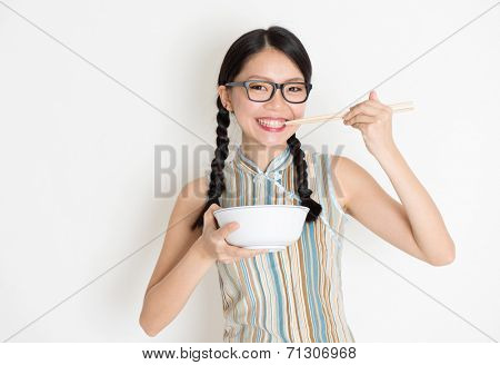 Portrait of Asian Chinese female eating, using chopsticks holding rice bowl, in retro revival style cheongsam standing on plain background.