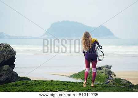 Long-haired surfer girl watching waves standing on the beach