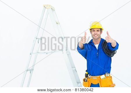 Portrait of happy electrician gesturing thumbs up while standing by ladder over white background
