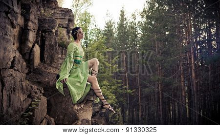 Elf Woman Is Sitting On The Rocks. Forest On The Background.