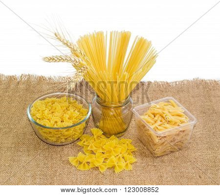 Uncooked dried long pasta and three wheat spikes in jar pasta several varieties and shapes in transparent tray and bowl and next to it on sackcloth on a light background
