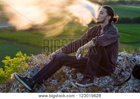 Handsome young man sitting on a rocks and smoking electronic cigarette at outdoors.