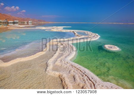 The path from salt picturesquely curls in salty water. Hotels are reflected in smooth water ashore. Israeli coast of the Dead Sea