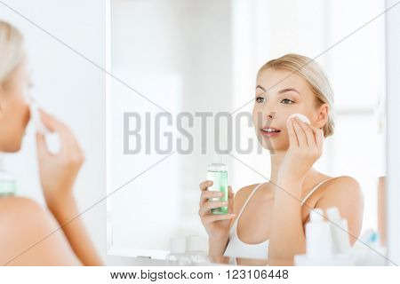 beauty, skin care and people concept - smiling young woman applying lotion to cotton disc for washing her face at bathroom