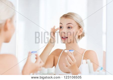 beauty, vision, eyesight, ophthalmology and people concept - young woman putting on contact lenses at mirror in home bathroom