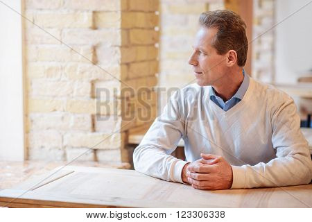 Let me think. Relaxed pleasant man holding hands in fists and looking in the window while sitting at the table