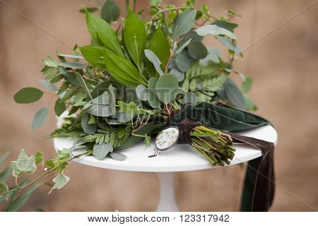 Green wedding bouquet of eucalyptus ivy fern aspidistra ruskus date fruit pink ranunkulyus with flax box green and brown velvet ribbons on white stand and brown on beige background
