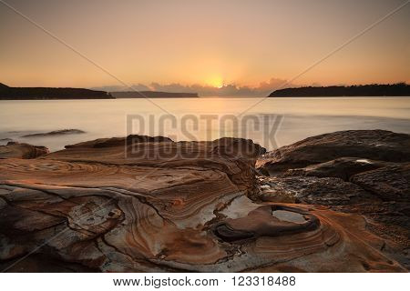 Sunrise from the pretty sandstone swirled rocks at Edwards Beach Balmoral with views east towards the Heads the entrance to Sydney Harbour as sunrays stream from behind clouds welcoming another new day.