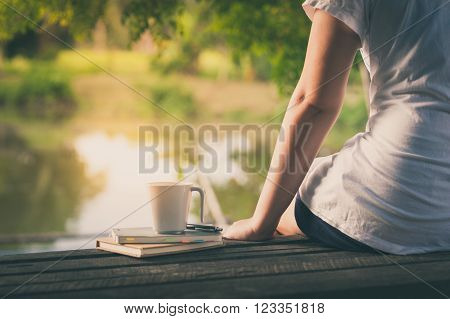 Young woman sitting and put down her hand beside coffee cup and notebooks on rustic wood bench with rural lake view in morning time on weekend