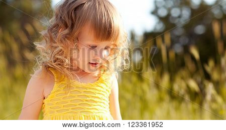 Kid girl 4-5 year old walking in meadow. Wearing casual yellow dress. Looking down. Childhood.