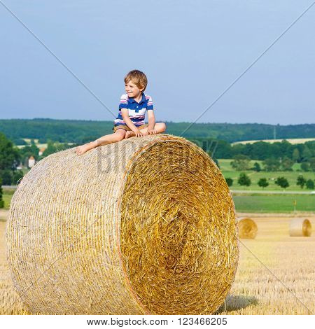 Happy little kid boy in traditional German bavarian clothes, leather shorts and check shirt. Child sitting on hay stack or bale and dreaming. Active outdoors leisure with children on warm summer day.