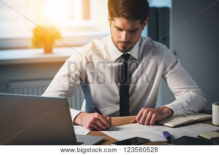Time to sign some contracts. Pensive young handsome man signing documents while sitting at his working place