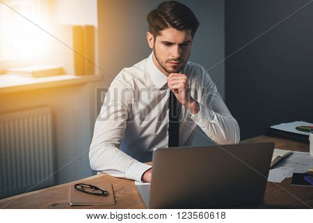 Totally concentrated. Pensive young handsome man using his laptop and keeping hand on chin while sitting at his working place