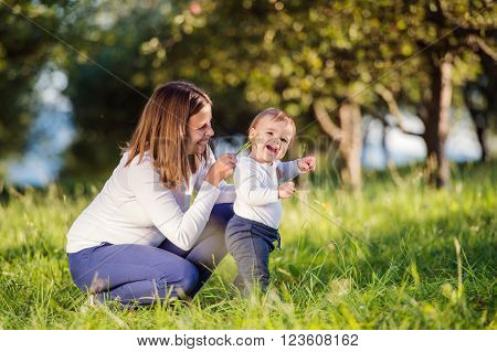 Mother holding her little son making first steps outside in green sunny nature, tickeling him with grass