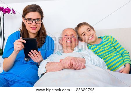 Kind doctor holding a digital tablet in her hand spending time with elderly woman when her grandson visits her.