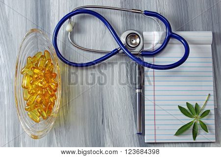 A blue stethoscope with a dish with tranparent yellow capsules and a notepad with a pen on a grey textured wooden background with copy space.
