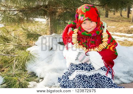 Child girl in Russian pavloposadskie folk scarf on head with floral print and with bunch of bagels on background of snow. Portrait of girl dressed in Russian style