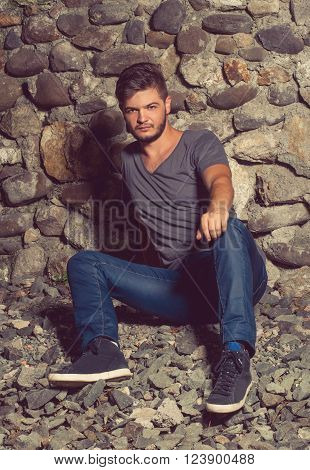 Young man wearing jeans and gray t-shirt sits down and posing in front of a stone wall