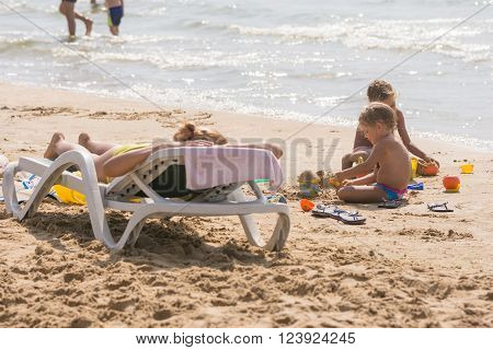 On The Sea Beach Near The Water With Sunbathing Chaise Longue Woman And Children Playing In The Sand