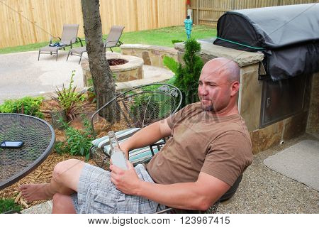 Cute bald muticultural man sitting on a patio in the back yard by bbq grill near his cell phone drinking a beer.