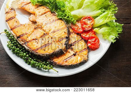 Grilled salmon steak served with grilled tomatoes, salad, lemon sliced, pepper seeds, rosemary, parsley on white plate on wooden table.