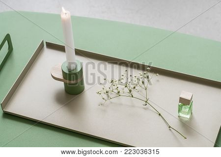 Metal-wooden green candlestick with burning candle on the beige metallic support on the green metal table in the studio. Next to it there is a white flower's branch and glass jar with wooden cover.