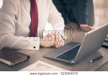 Team lead in white shirt and red tie and manager in suit discuss data on laptop.