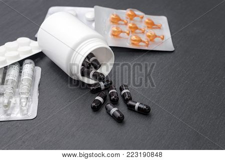 Supplement capsules are poured from white bottle, tablets and ampoules on slate board. Medicine theme, health care concept