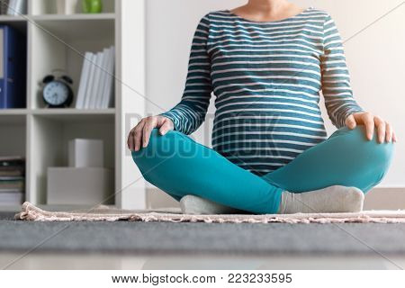Pregnant woman practicing yoga exercise at home. Pregnancy yoga and indoors fitness concept. Ungraded low contrast footage with natural window light.