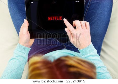 Benon, France - January 21, 2018: woman sitting cross-legged on her couch and using her touch pad to connect to the Netflix app. Netflix Inc. is an American multinational entertainment company founded on 1997