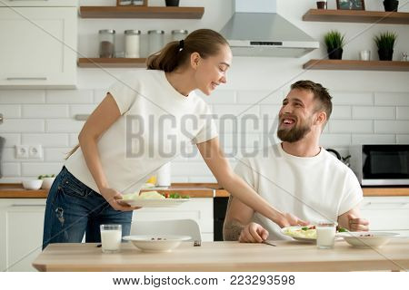 Attractive caring wife serving food cooked for husband in cozy kitchen, grateful man appreciating woman prepared healthy breakfast dinner, happy couple enjoying meal together at home dining table