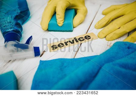Blue cleaning supplies: sponges, brushes, windex, hands in rubber gloves washing the surface, service inscription, white wooden background