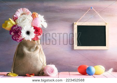 Flower bouquet with painted eggs and a chalkboard - Easter banner with a bouquet of flowers in a jute sack, painted eggs and a blank blackboard hanging on a purple wooden background, on a sunny day.