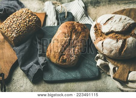 Various bread selection. Rye, wheat and multigrain rustic bread loaves on kitchen towels and wooden boards over grey concrete stone background