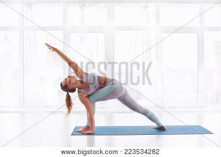 Beautiful young woman practices yoga asana Parivritta Parshvakonasana - Revolving side angle posture at the bright yoga class with large windows