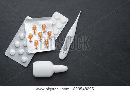 Thermometer, nasal drops and tablets on dark slate board. Flat lay. Meds for flue treatment, health care concept
