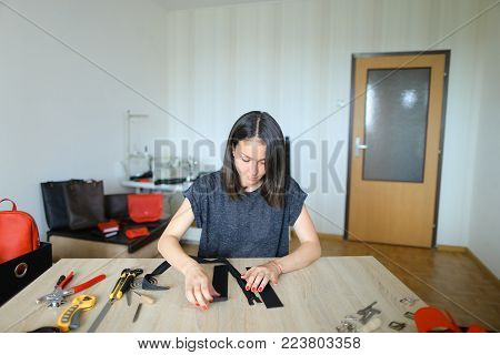 female fixing snap on leather pattern using special mechanism. Young designer decided to try to make women bags for sale. Black-haired girl with red nails wearing grey T-shirt sitting near table in workshop. Concept of creative work, own business or handw