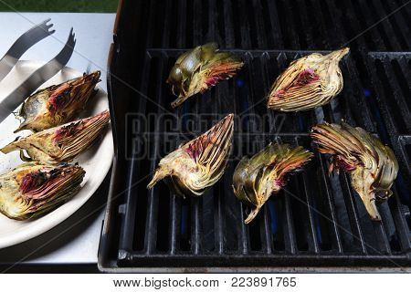 Grilled Artichokes: Artichoke halves on a gas grill. A plate of done vegetables is on the side tray. 