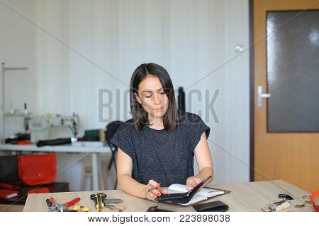 female fixing snap on leather pattern using special mechanism. Young designer decided to try to make women bags for sale. Black-haired girl with red nails wearing grey T-shirt sitting near table in workshop. Concept of creative work, own business or handw
