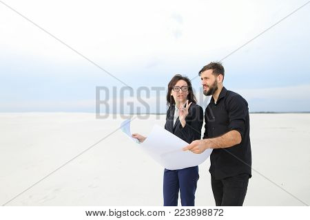 CEO male and CFO female holding whatman paper, colleagues discuss project of wind power plant in deserted place. Young woman and bearded guy standing among sands looking around. Concept of exploring new territories and designing or alternative energy sour
