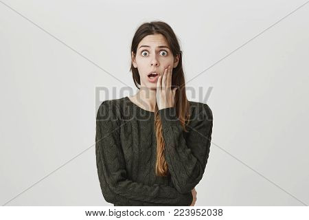 Frightened scared dark-haired woman wears green sweater, looks with terrified expression, being afraid of something, expresses negative emotions and feelings, poses against gray background.
