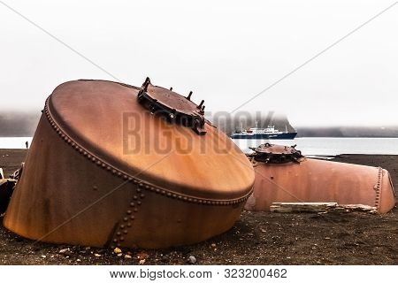 Abandoned Norwegian Whale Hunter Station Rusty Blubber Tanks With Cruise Vessel In The Bay , At Dece