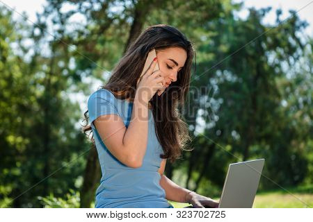 Young Brunette Srudent In A Blue Dress Sitting With A Laptop In A Park And Talking On The Phone. Con