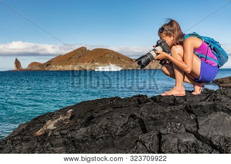 Galapagos tourist photographing marine iguana on Santiago Island in Galapagos Islands. Cruise ship and Pinnacle Rock and Bartolome Island in background. Famous Galapagos cruise ship tour destination.