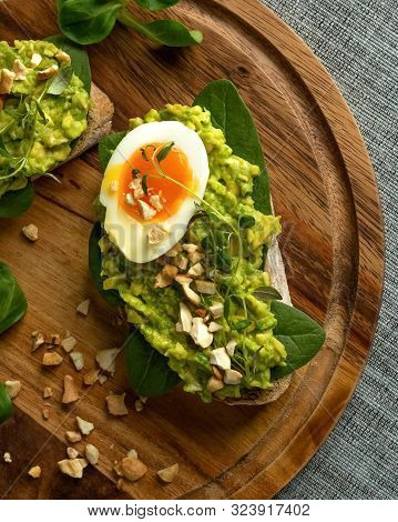 Close Up Of An Avocado Toasts With Soft Boiled Egg, Herbs, Spinach, Salad And Nuts On A Round Wooden