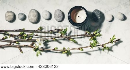 Flat lay composition with beautiful spring cherry branches, natural candle and grey stones on white marble background. SPA, yoga, relaxation, natural cosmetics, zen like concept