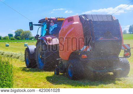 A Red Tractor And A Hay Baler At Work In A Rural Field In Making Hay. Back View.