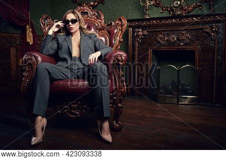 Wealthy lifestyle. Sexual woman in fashionable pantsuit and black sunglasses alluring in a vintage armchair in a luxury apartments. Fashion shot.