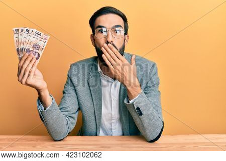 Young hispanic man holding mexican pesos sitting on the table covering mouth with hand, shocked and afraid for mistake. surprised expression 