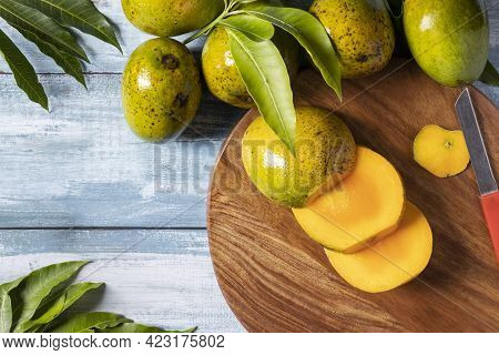 Top View Of Ripe Mango Slices On Wooden Chopping Board Along With Mangoes And Leaves On Wooden Table